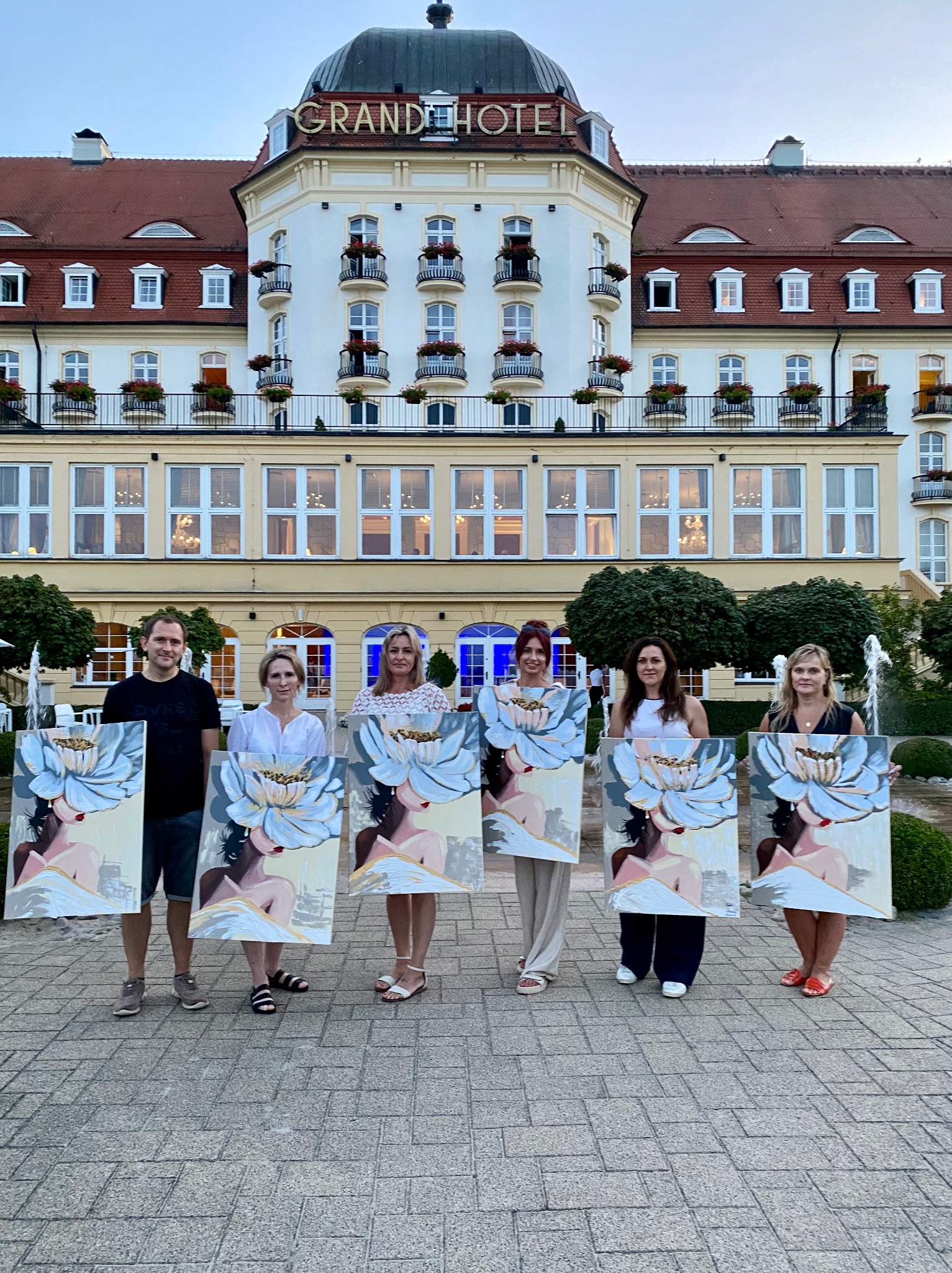 Grupa osób prezentuje obrazy z motywem kwiatowym na tle Grand Hotelu, prawdopodobnie po warsztatach artystycznych zorganizowanych przez agencję eventową.