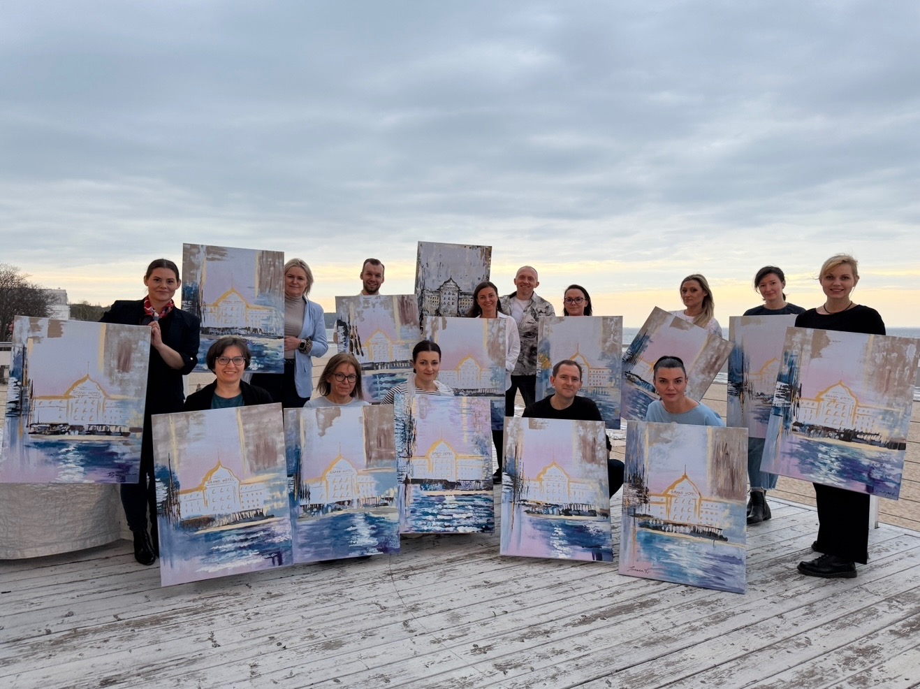 Grupa osób prezentuje obrazy z motywem Grand Hotelu na tle pochmurnego nieba i drewnianego podestu, prawdopodobnie po warsztatach malarskich.