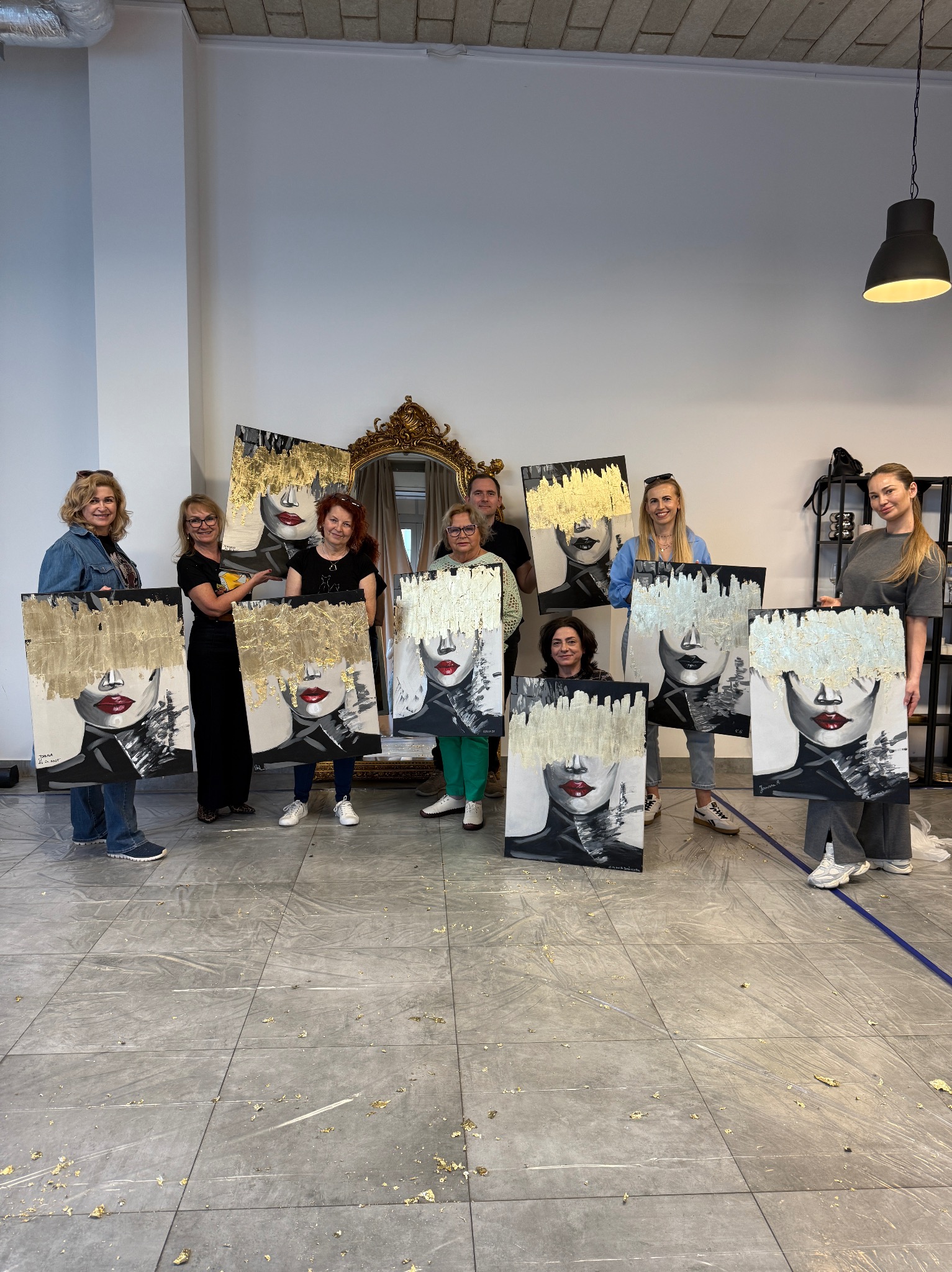 Grupa osób prezentuje obrazy z abstrakcyjnymi portretami w odcieniach szarości z elementami złota, po warsztatach artystycznych. Złote płatki na podłodze.
