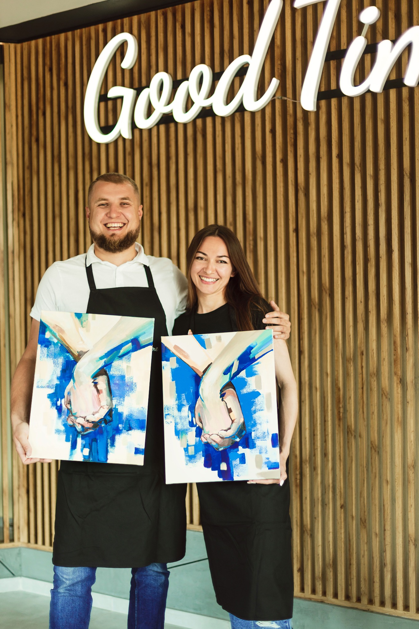 Dwoje uśmiechniętych artystów prezentuje obrazy z motywem dłoni na tle drewnianej ściany z napisem „Good Times”. Warsztaty artystyczne.