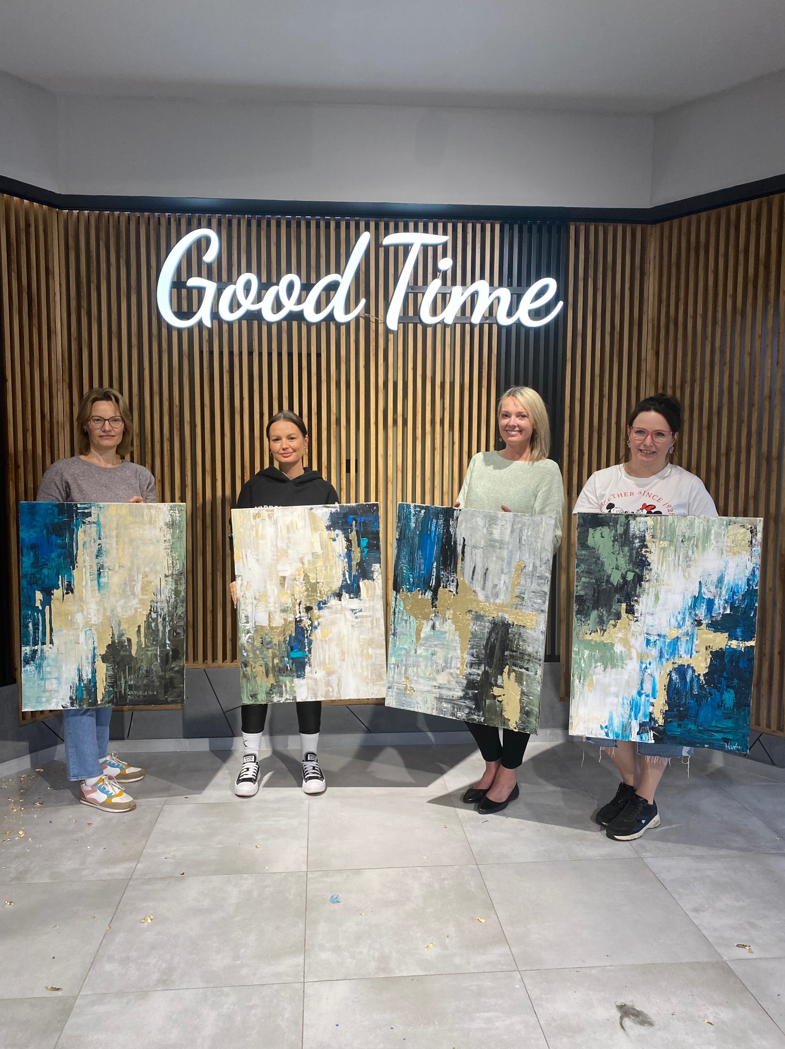 Cztery kobiety prezentują abstrakcyjne obrazy z akcentami złota na tle drewnianej ściany z neonem 'Good Time', widoczne drobinki złota na podłodze.