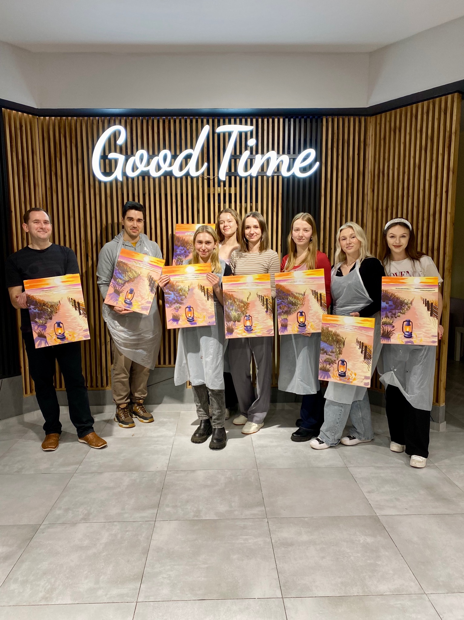 Grupa osób prezentuje obrazy z motywem latarni morskiej, efekt warsztatów malarskich. W tle dekoracyjna ściana z napisem neonowym 'Good Time'.