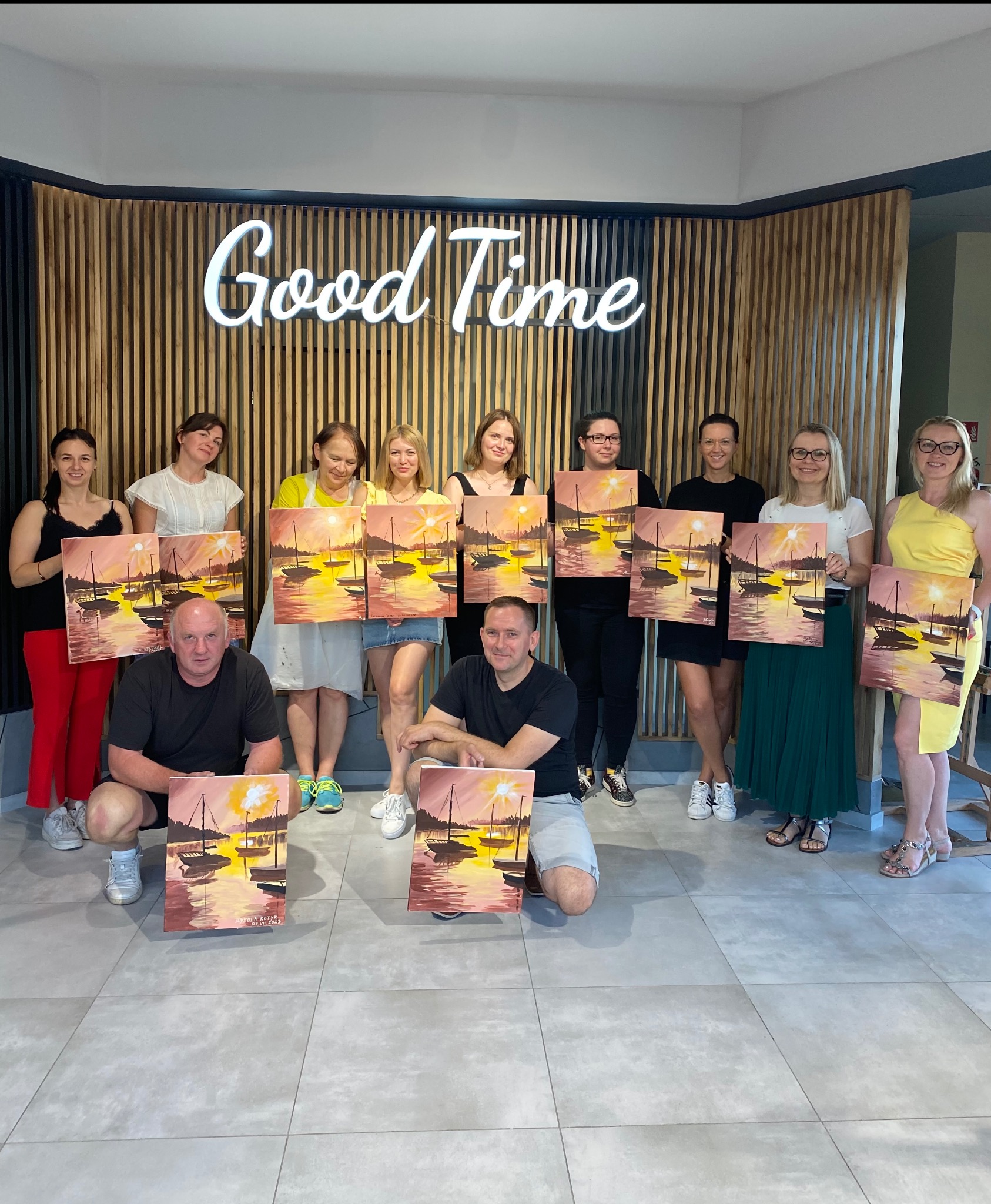 Grupa osób prezentuje obrazy z motywem zachodu słońca nad wodą, wykonane podczas warsztatów malarskich. W tle dekoracyjna ściana z napisem 'Good Time'.