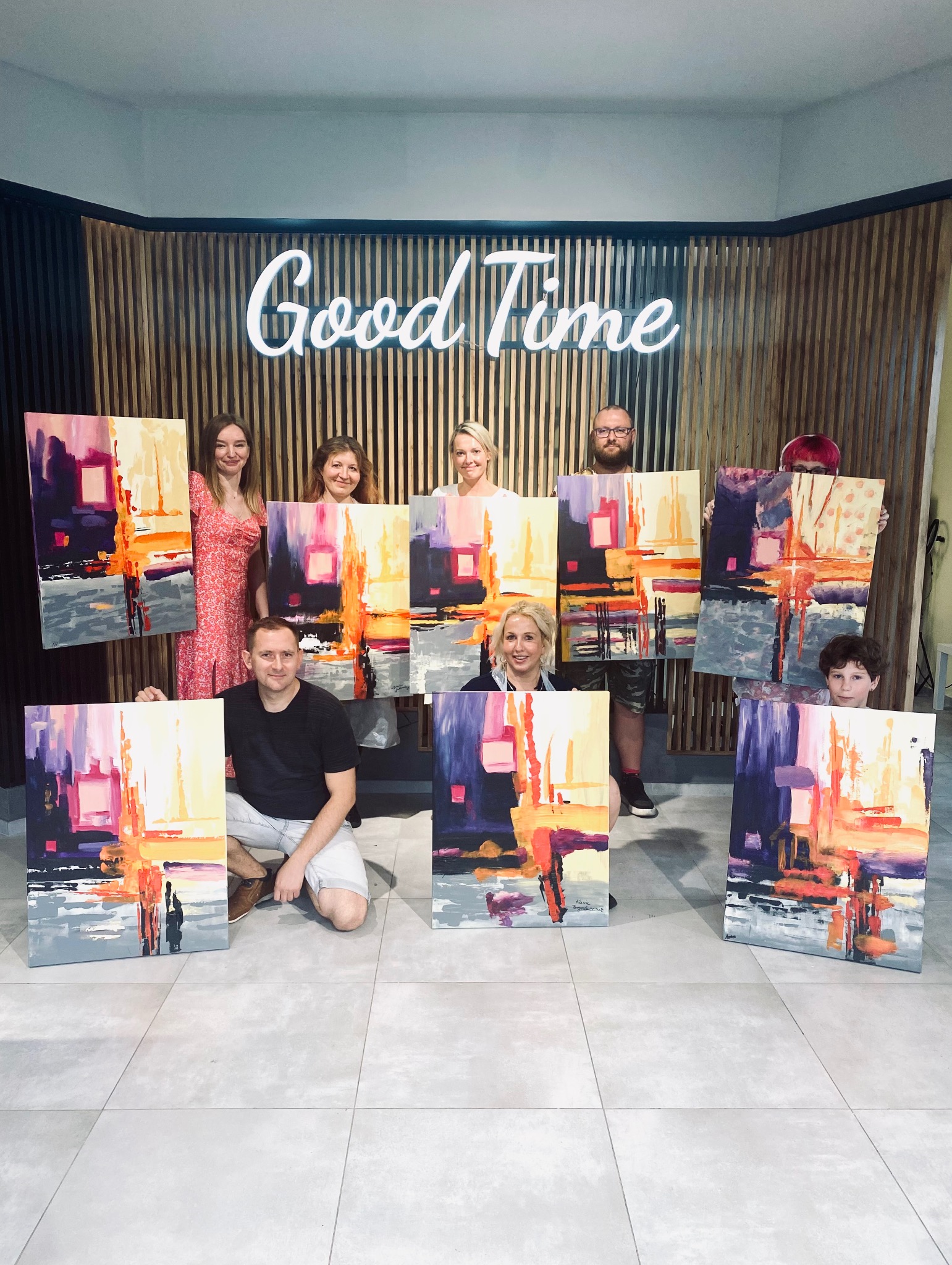 Grupa osób prezentuje abstrakcyjne obrazy na płótnach, z neonowym napisem 'Good Time' w tle, sugerującym artystyczne warsztaty lub event.