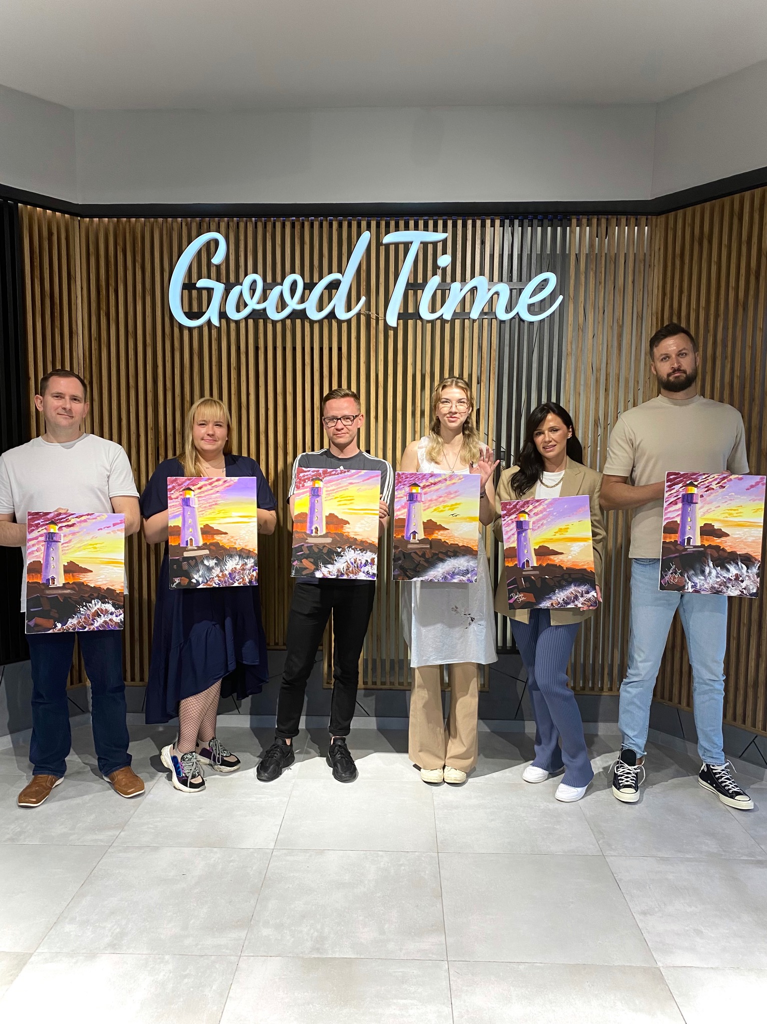 Sześć osób prezentuje obrazy z motywem latarni morskiej na tle drewnianej ściany z neonowym napisem 'Good Time'.