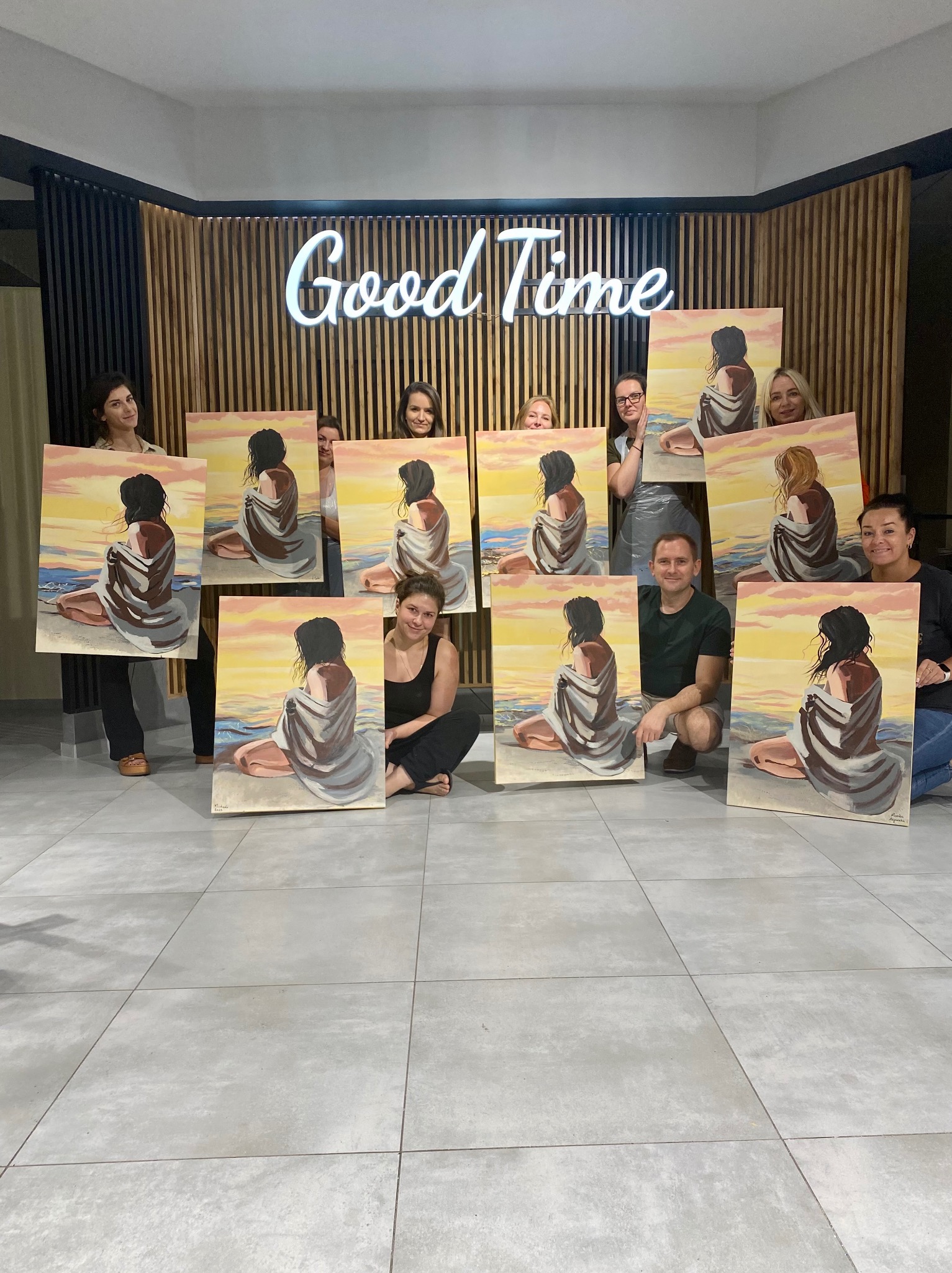 Grupa osób prezentuje obrazy z motywem kobiety na plaży, wykonane podczas warsztatów malarskich w studio z neonowym napisem 'Good Time' w tle.