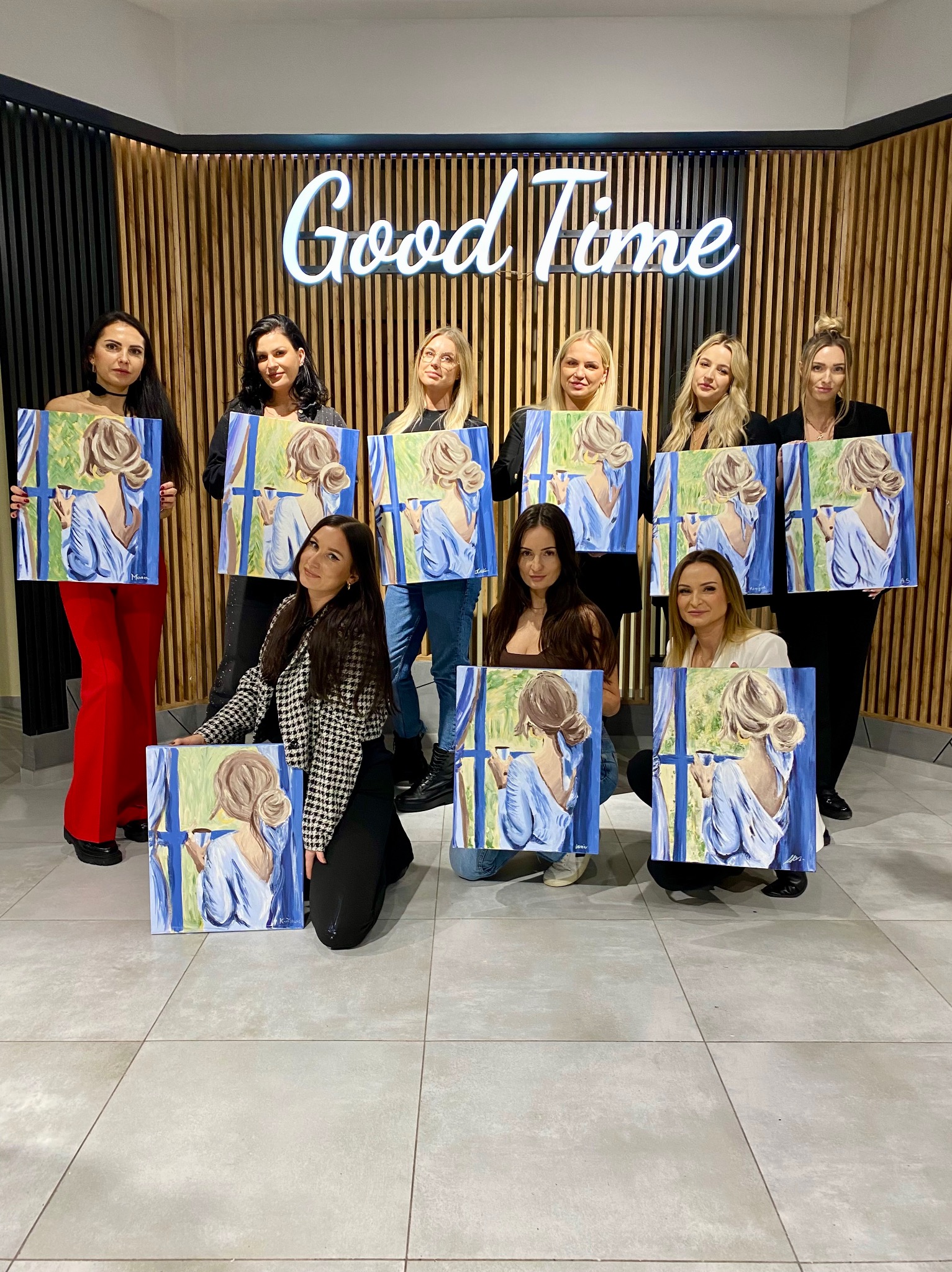 Grupa kobiet prezentuje obrazy z motywem kobiety z filiżanką na tle okna, w tle neon z napisem 'Good Time', sugerujący warsztaty malarskie.