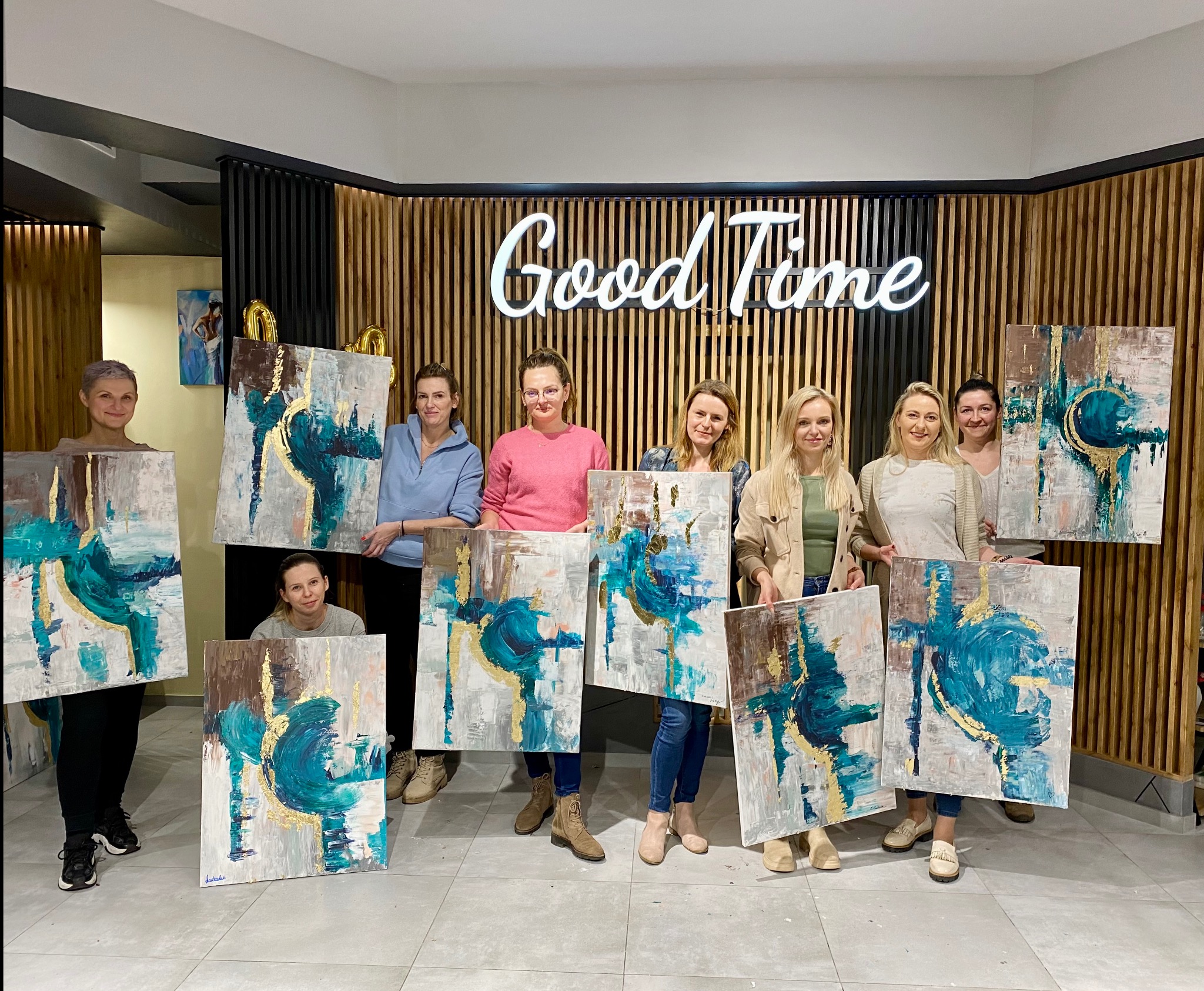 Warsztaty malarskie w Gdańsku: grupa kobiet prezentuje swoje abstrakcyjne obrazy z akcentami złota na tle drewnianej ściany z napisem 'Good Time'.