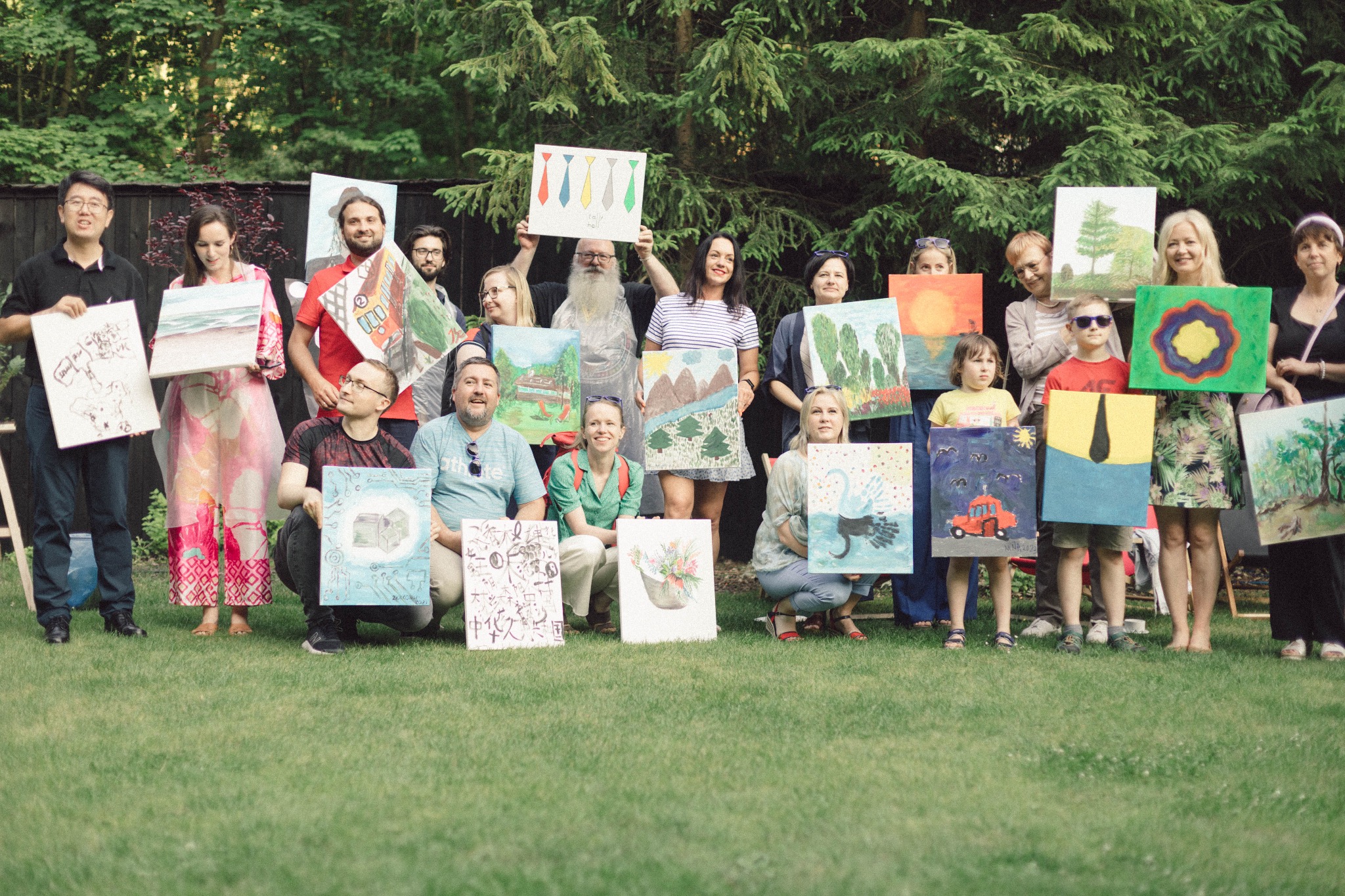 Grupa osób prezentuje różnorodne obrazy na płótnach podczas plenerowego wydarzenia artystycznego, na tle zieleni.