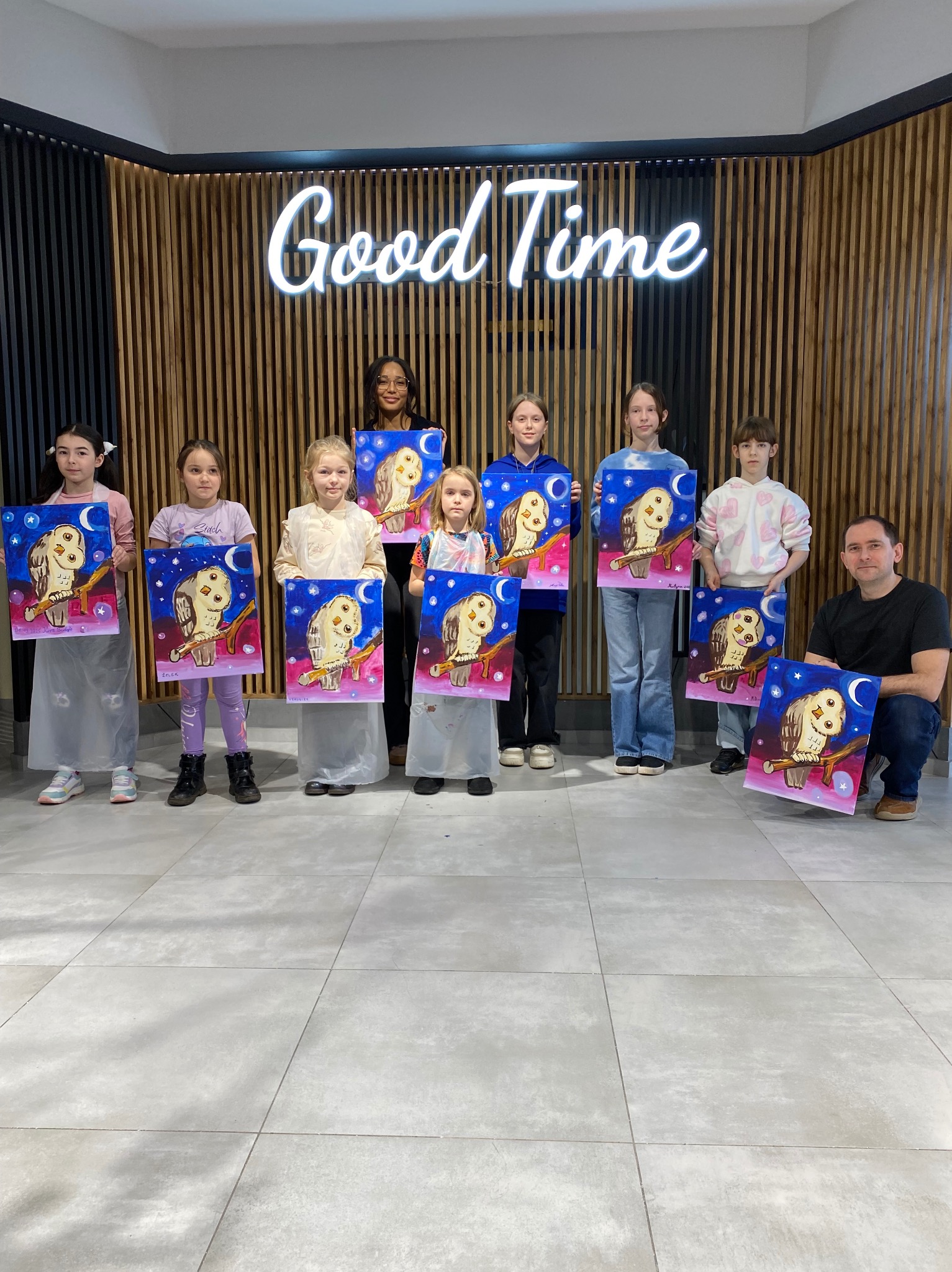 Grupa dzieci i dwoje dorosłych prezentuje obrazy sowy na tle ściany z napisem 'Good Time' w jasnym wnętrzu. Uczestnicy warsztatów artystycznych trzymają swoje prace.