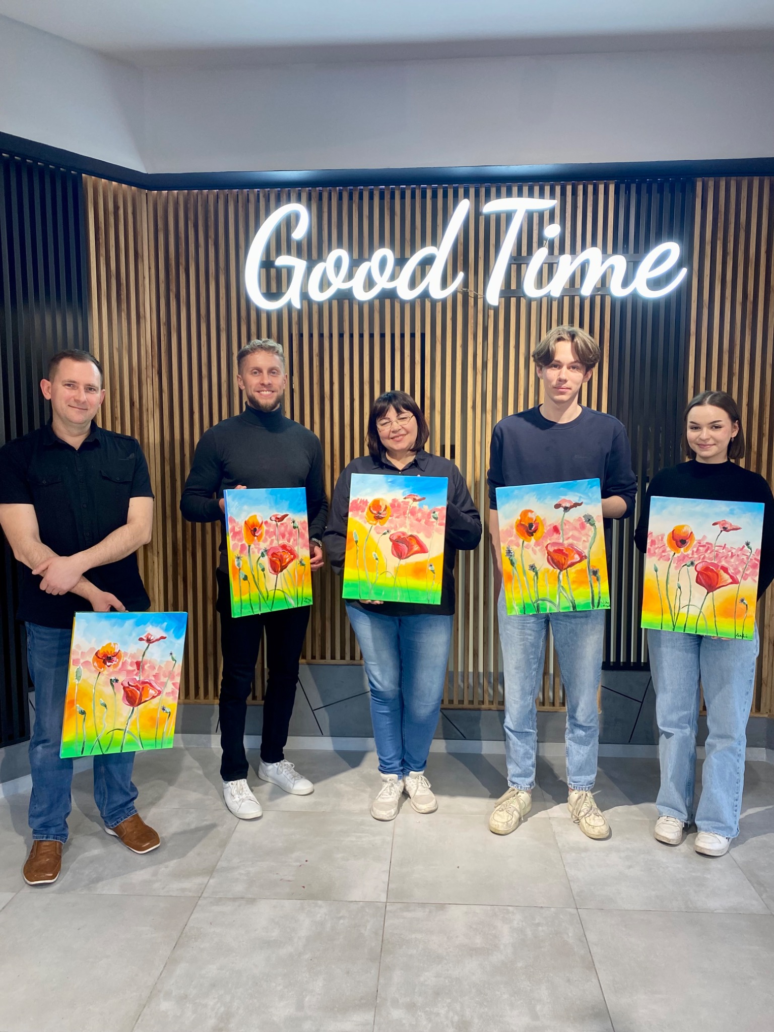 Grupa osób prezentuje akwarelowe obrazy z motywem maków na tle drewnianej ściany z neonowym napisem 'Good Time'. Każdy obraz ma inne tło kolorystyczne.
