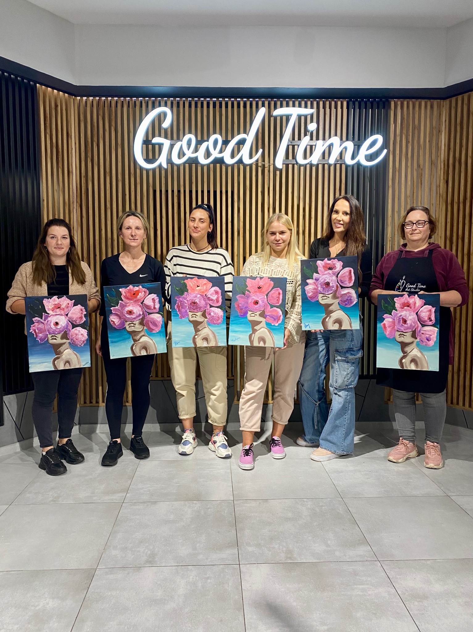 Grupa sześciu kobiet prezentuje obrazy z motywem kwiatowym na tle ściany z neonem 'Good Time', uchwycone w jasnym, przestronnym wnętrzu studia artystycznego.