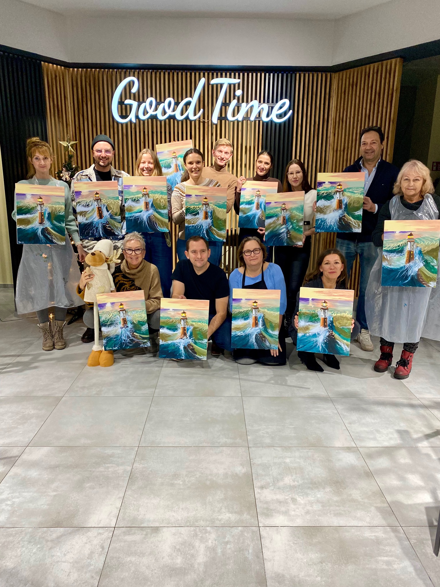 Grupa osób prezentuje obrazy z motywem latarni morskiej, wykonane podczas warsztatów malarskich. W tle neonowy napis 'Good Time' i dekoracja z gwiazdą.