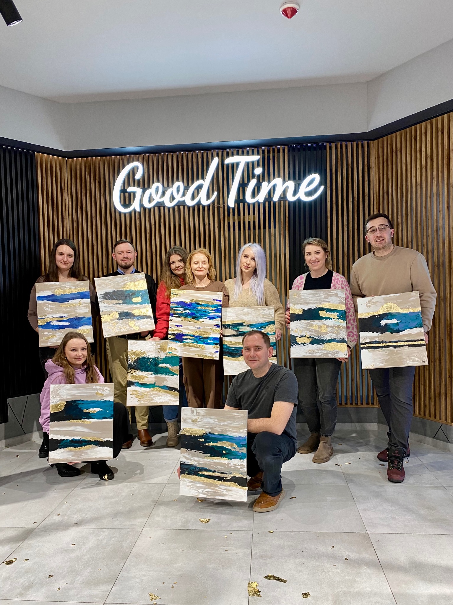Grupa osób prezentuje abstrakcyjne obrazy z elementami złota na tle ściany z neonem 'Good Time', co sugeruje warsztaty artystyczne.