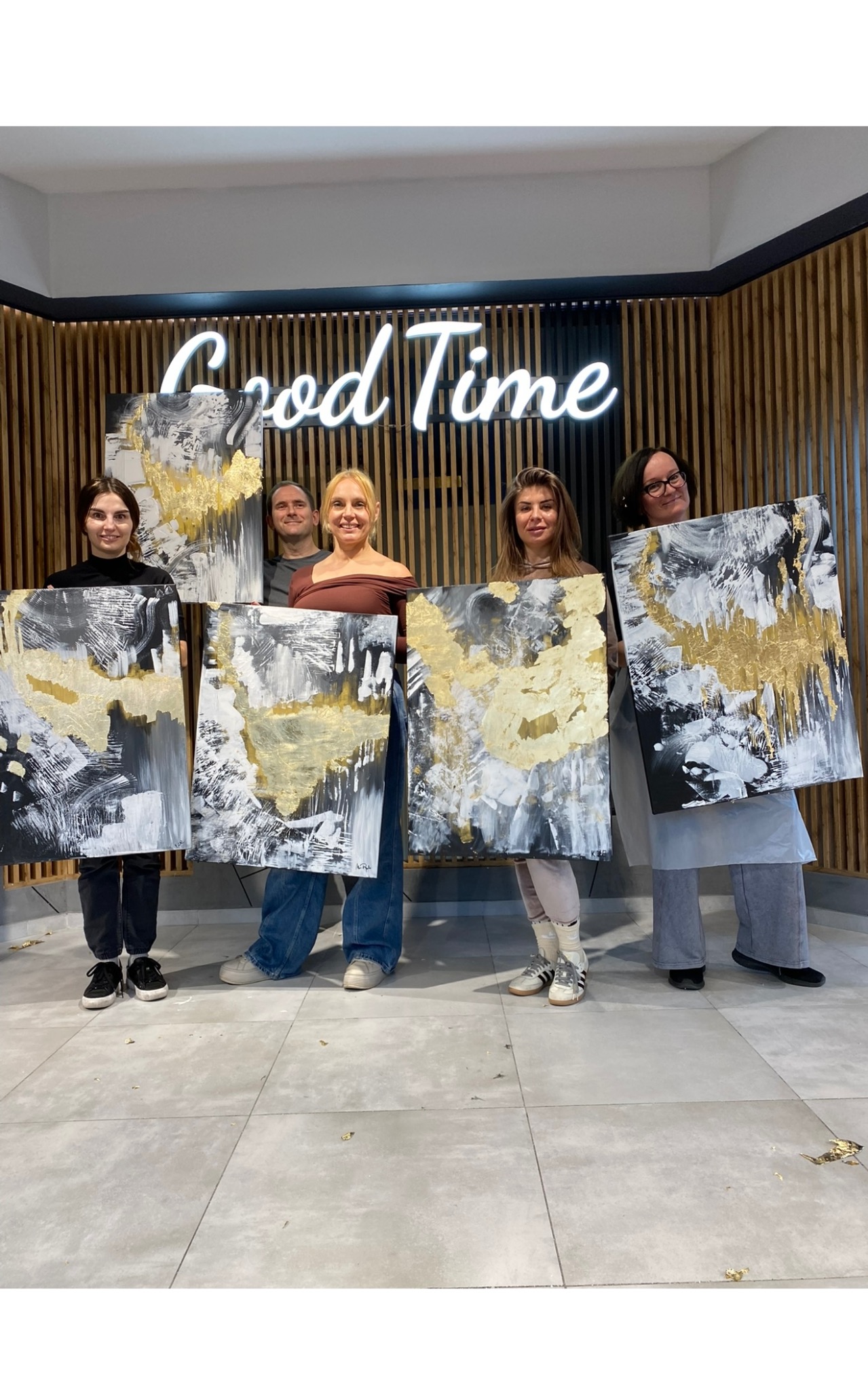 Grupa osób prezentuje abstrakcyjne obrazy z elementami złota na tle neonowego napisu 'Good Time' i drewnianej ściany. Podłoga pokryta jest drobnymi kawałkami złota.