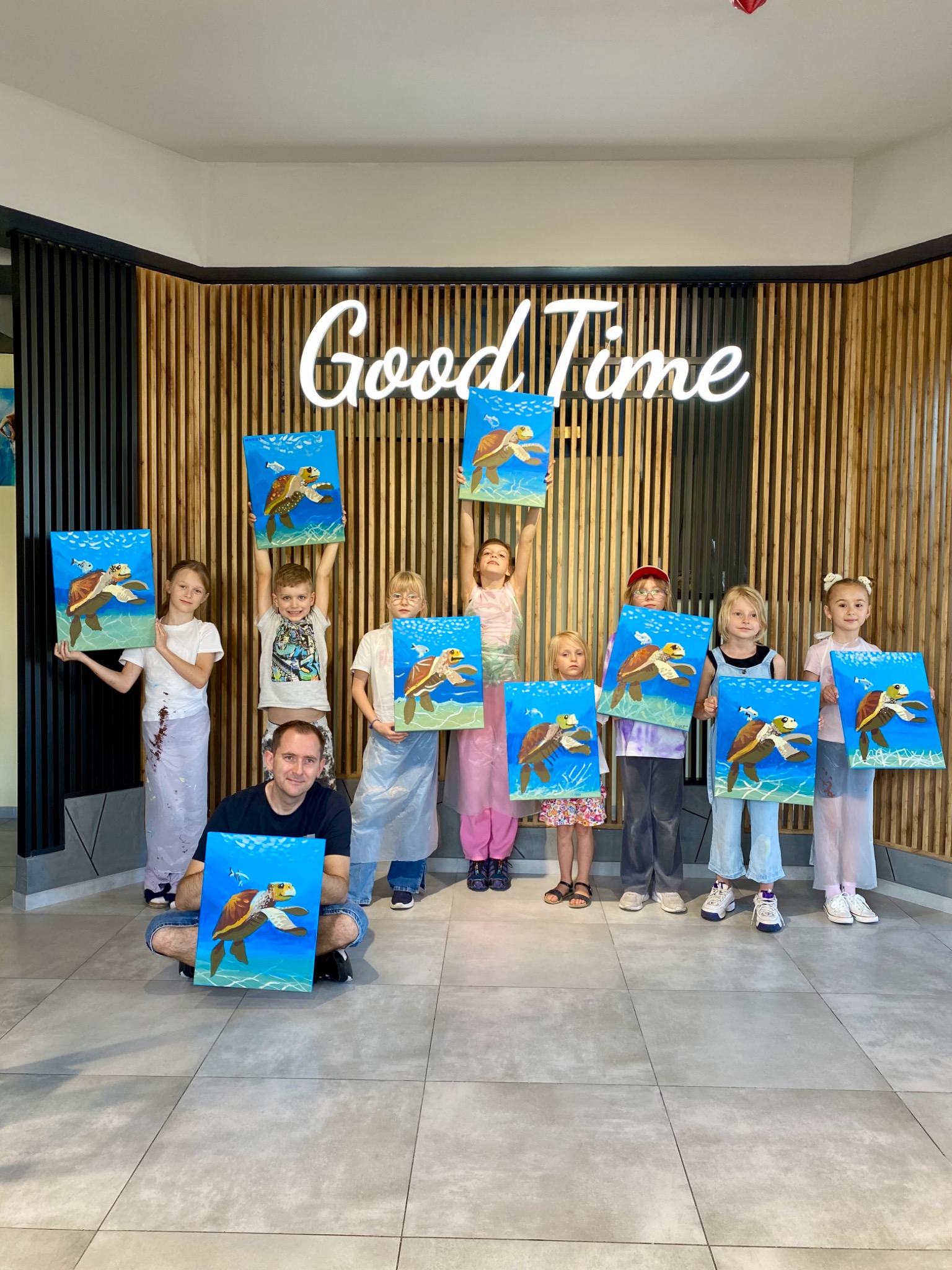 Grupa dzieci z instruktorem prezentuje obrazy żółwi morskich na płótnach, wykonane podczas warsztatów malarskich w jasnym wnętrzu z neonem 'Good Time'.