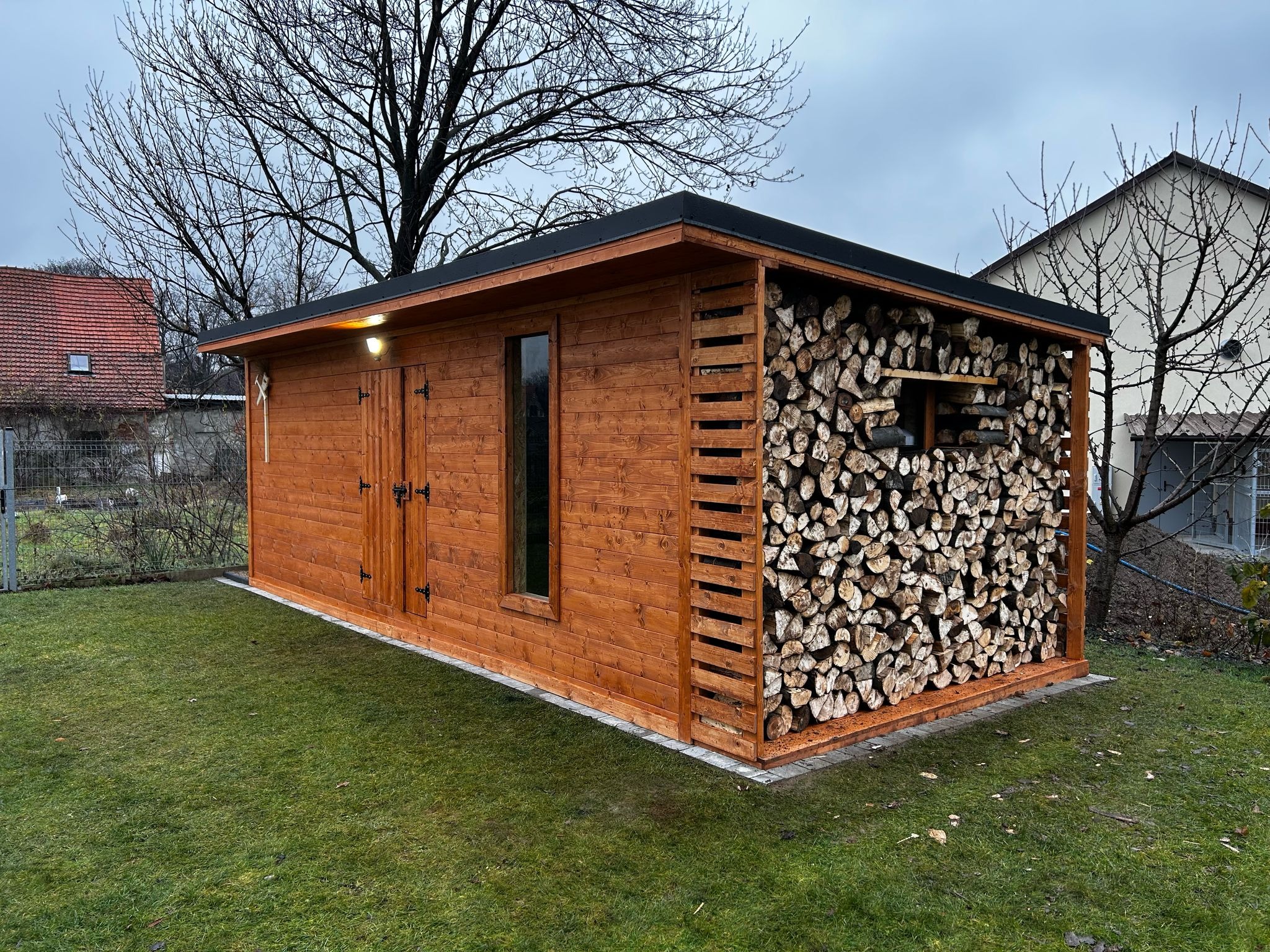 Funkcjonalny, drewniany domek narzędziowy z miejscem na składowanie drewna, na tle zielonego trawnika i zabudowań. Widoczne drzwi, okno i konstrukcja z drewna.
