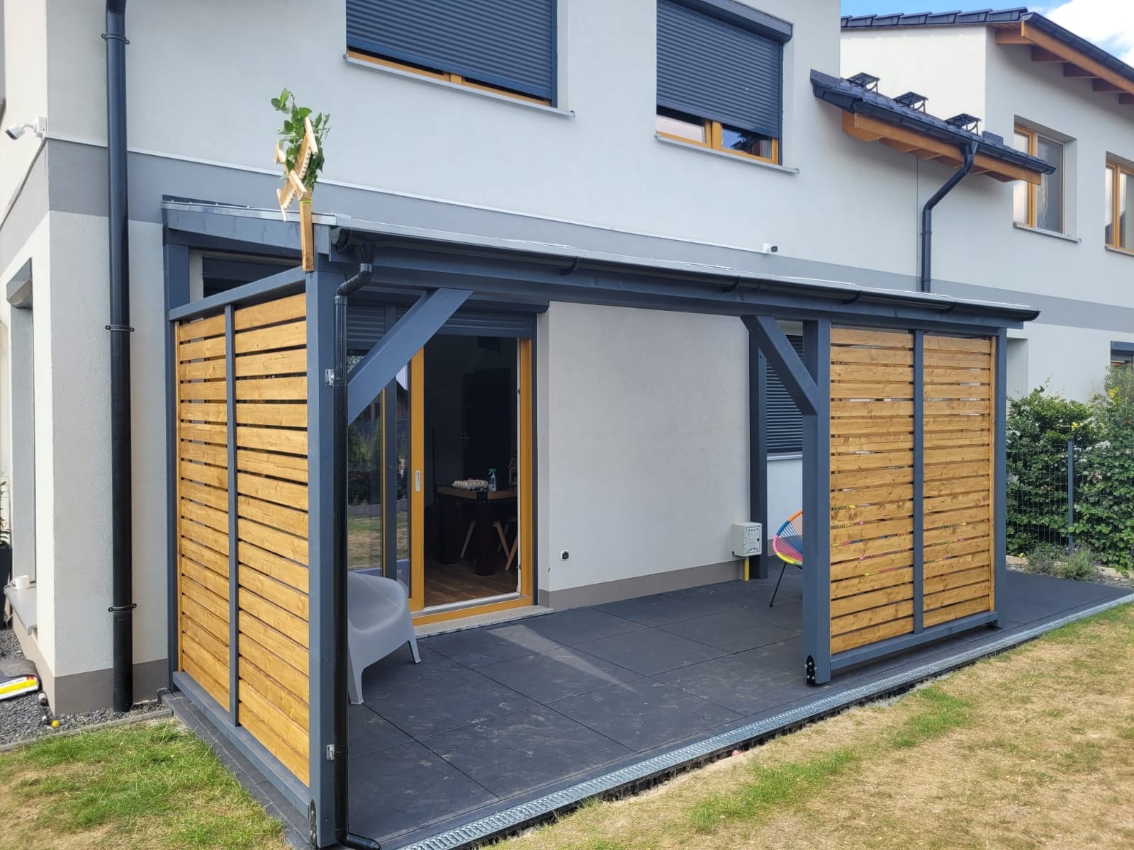 Nowoczesny taras z zadaszeniem i drewnianą przesłoną. Szare elementy konstrukcyjne, płytki w ciemnym kolorze, widoczne detale wykończenia. Całość wkomponowana w otoczenie domu.