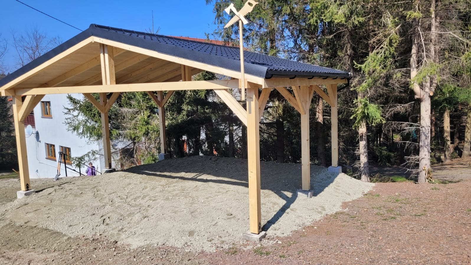 Surowa, drewniana konstrukcja z czarnym dachem, prawdopodobnie altana, na tle drzew i budynku. Na szczycie konstrukcji ozdobny wiatrowskaz z motywem piły i siekiery.