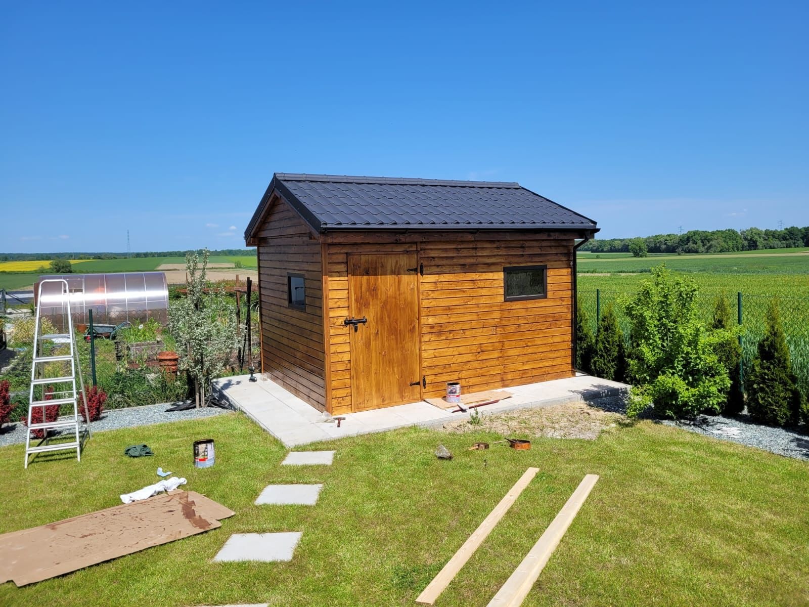 Drewniana altana z ciemnym dachem na betonowej podstawie, otoczona zielonym trawnikiem i polami uprawnymi w słoneczny dzień. Widoczne narzędzia i materiały budowlane.