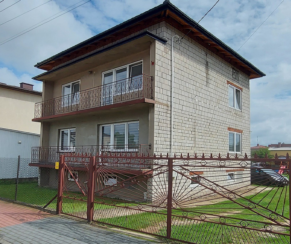 Dwupiętrowy dom z pustaków z balkonami i dekoracyjną, metalową bramą w kolorze czerwonej cegły, widziany z poziomu chodnika.