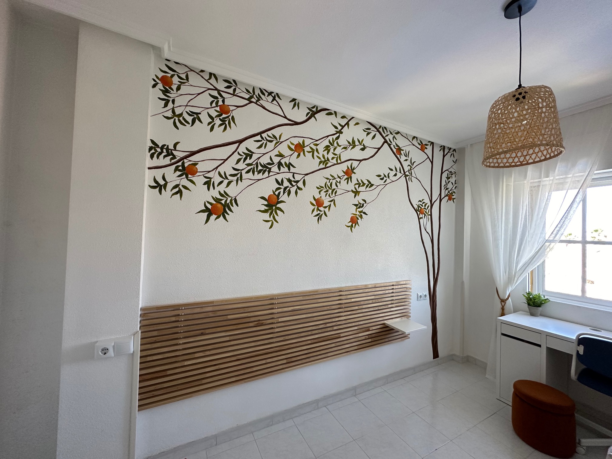 Akrylowa malowanka na ścianie w pokoju: drzewo pomarańczowe rozciąga się nad drewnianym zagłówkiem. W tle biurko i okno z firanką.