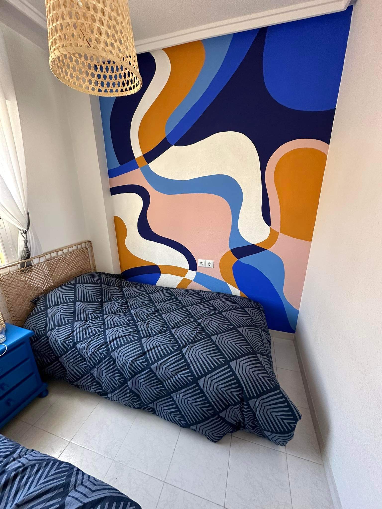 Pokój z łóżkiem i abstrakcyjnym malowidłem ściennym w odcieniach błękitu, różu, bieli i pomarańczy. W rogu lampa z wikliny.