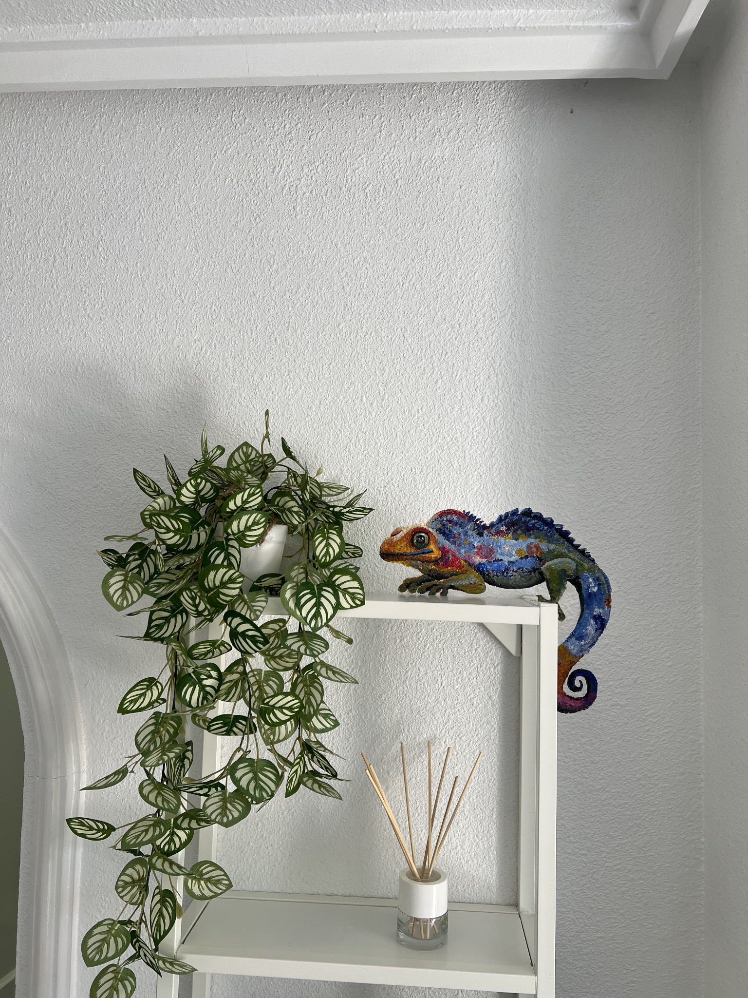 Kolorowa rzeźba kameleona, roślina doniczkowa i dyfuzor zapachowy na białym regale, na tle białej ściany o nierównej fakturze.