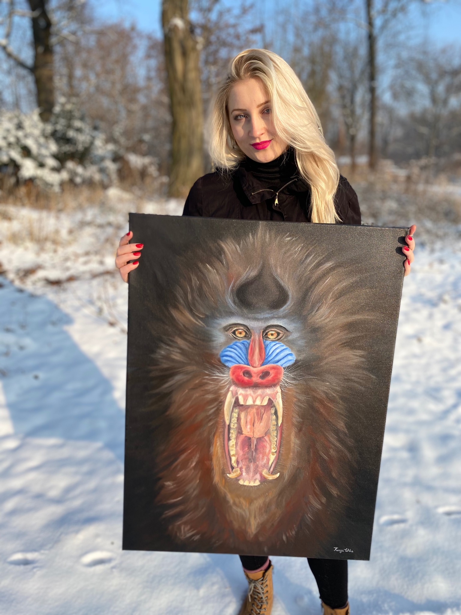 Blondynka prezentuje obraz olejny mandryla na tle zimowego krajobrazu. Intensywne kolory malowidła kontrastują z bielą śniegu. Widoczny podpis artysty w prawym dolnym rogu.