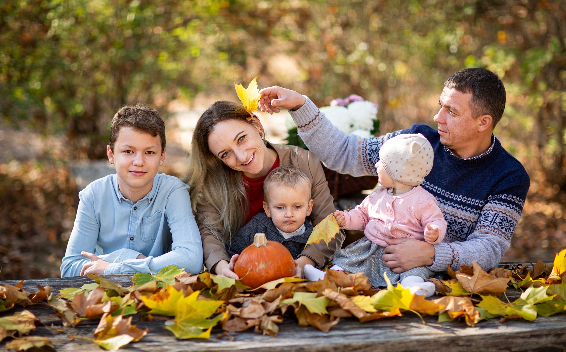 Jesienna sesja rodzinna: rodzice z trójką dzieci siedzą na ławce, otoczeni kolorowymi liśćmi. Ojciec wkłada liść za ucho mamy, obok dynia i kwiaty w koszu.