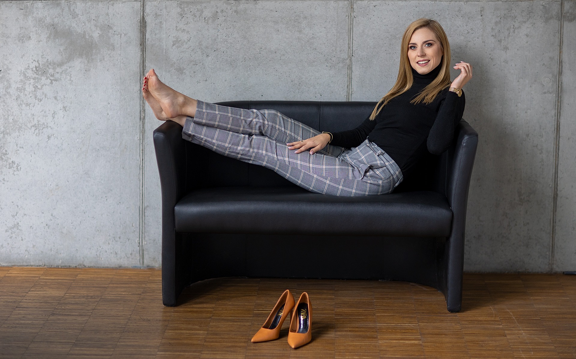 Kobieta w czarnym golfie i spodniach w kratę relaksuje się na czarnej sofie, obok leżą szpilki. Zdjęcie biznesowe w nowoczesnym wnętrzu na tle betonowej ściany.