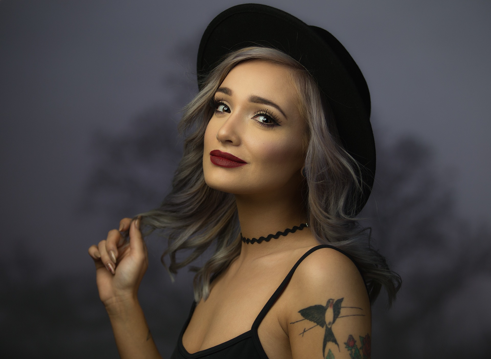 Portret kobiety w czarnym kapeluszu, z tatuażem na ramieniu, na rozmytym tle. Uśmiech, choker i srebrne włosy dodają charakteru.