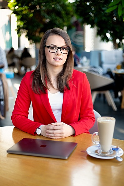 Kobieta w czerwonej marynarce siedzi przy stole w kawiarni z laptopem i kawą latte. Profesjonalny wizerunek biznesowy, naturalne światło, delikatne rozmycie tła.