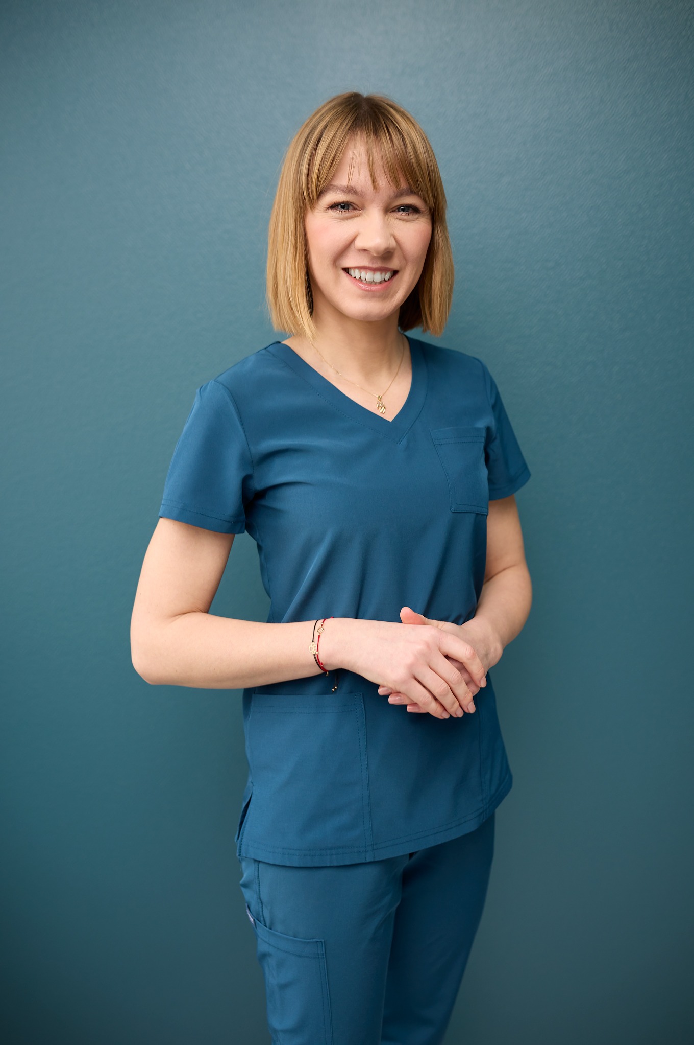 Portret uśmiechniętej kobiety w turkusowym uniformie medycznym na tle ściany w odcieniu petrol. Widoczne dłonie splecione na wysokości brzucha i złoty naszyjnik.