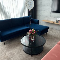 Brilans - Udekorowany salon z ciemnoniebieską sofą narożną, czarnym stolikiem z kwiatami i balonem. Konfetti w kształcie serc na stoliku. Stonowana kolorystyka wnętrza.