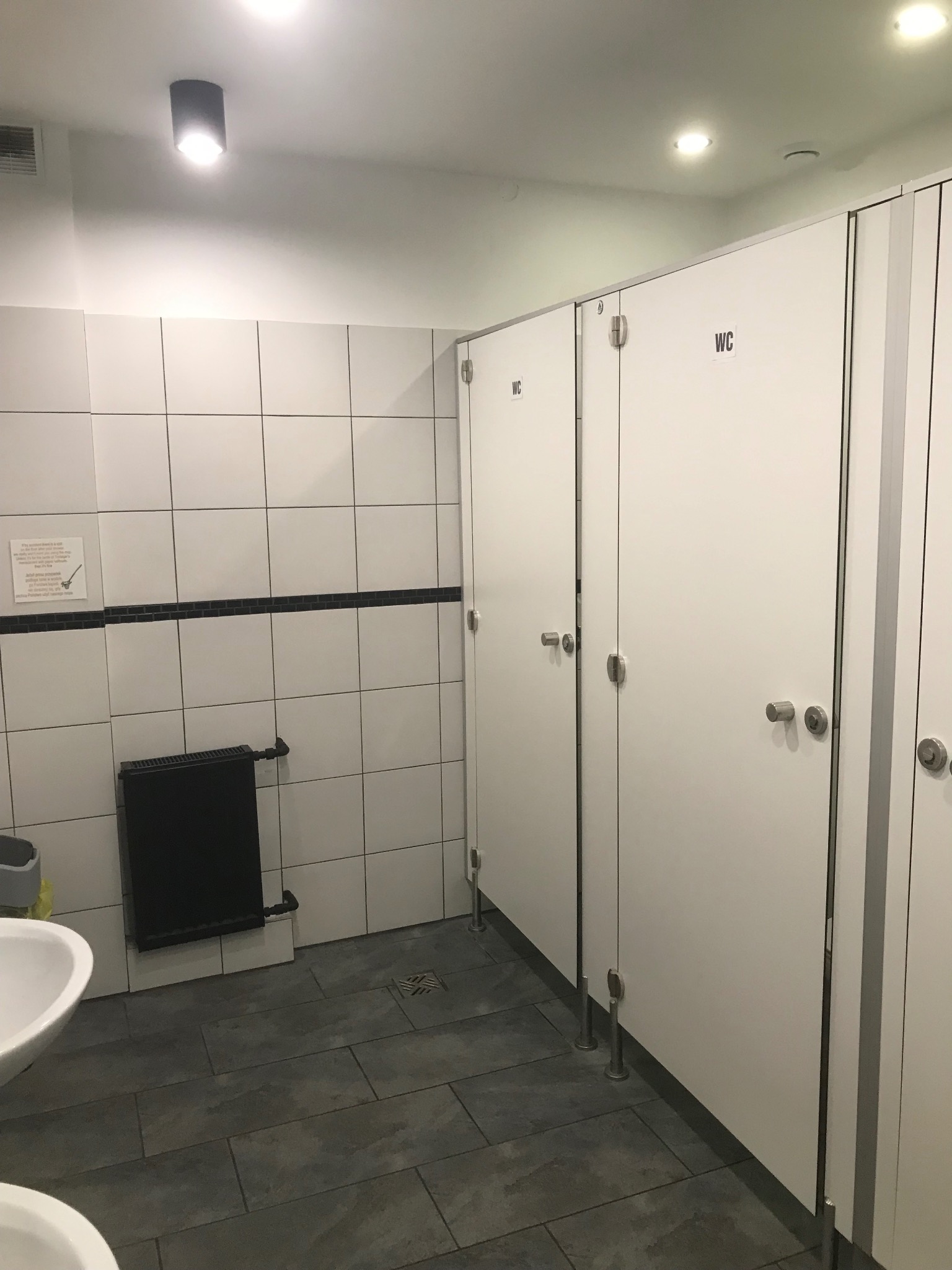 Czysta toaleta publiczna z białymi kabinami WC i szarymi płytkami podłogowymi. Widoczne umywalki i grzejnik. Oświetlenie sufitowe.