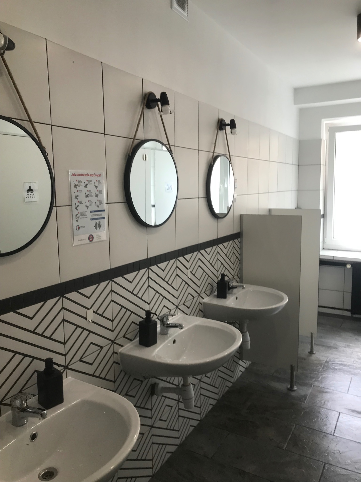Wnętrze umywalni publicznej z trzema umywalkami, okrągłymi lustrami na sznurkach i czarno-białymi kafelkami z geometrycznym wzorem na ścianach.