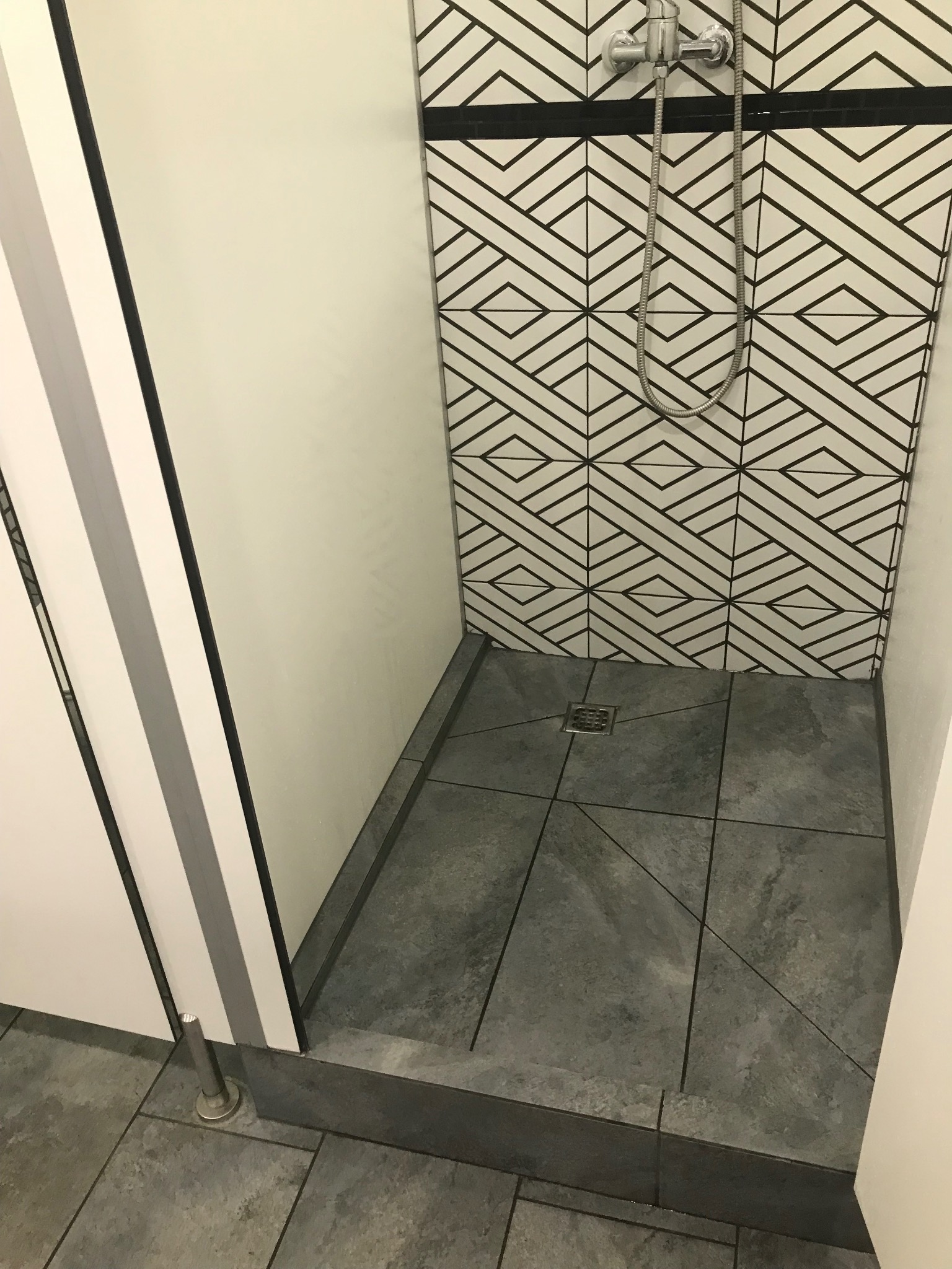 Nowoczesny prysznic z geometrycznymi płytkami na ścianie i szarymi płytkami na podłodze, widoczny odpływ liniowy i chromowana bateria.