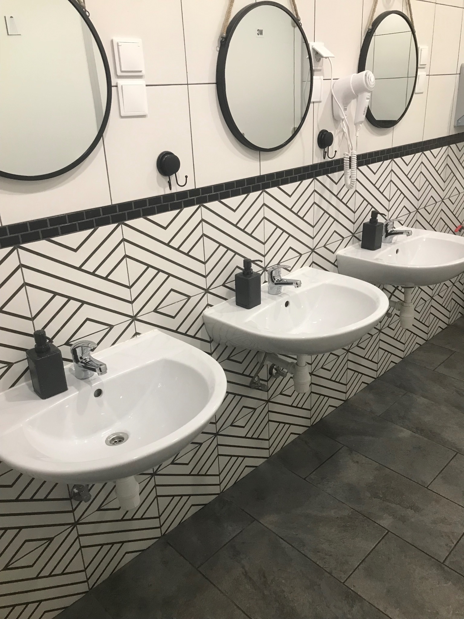 Rząd trzech białych umywalek z czarnymi dozownikami mydła, okrągłe lustra z czarną ramą i suszarka do włosów na ścianie z geometrycznym wzorem.