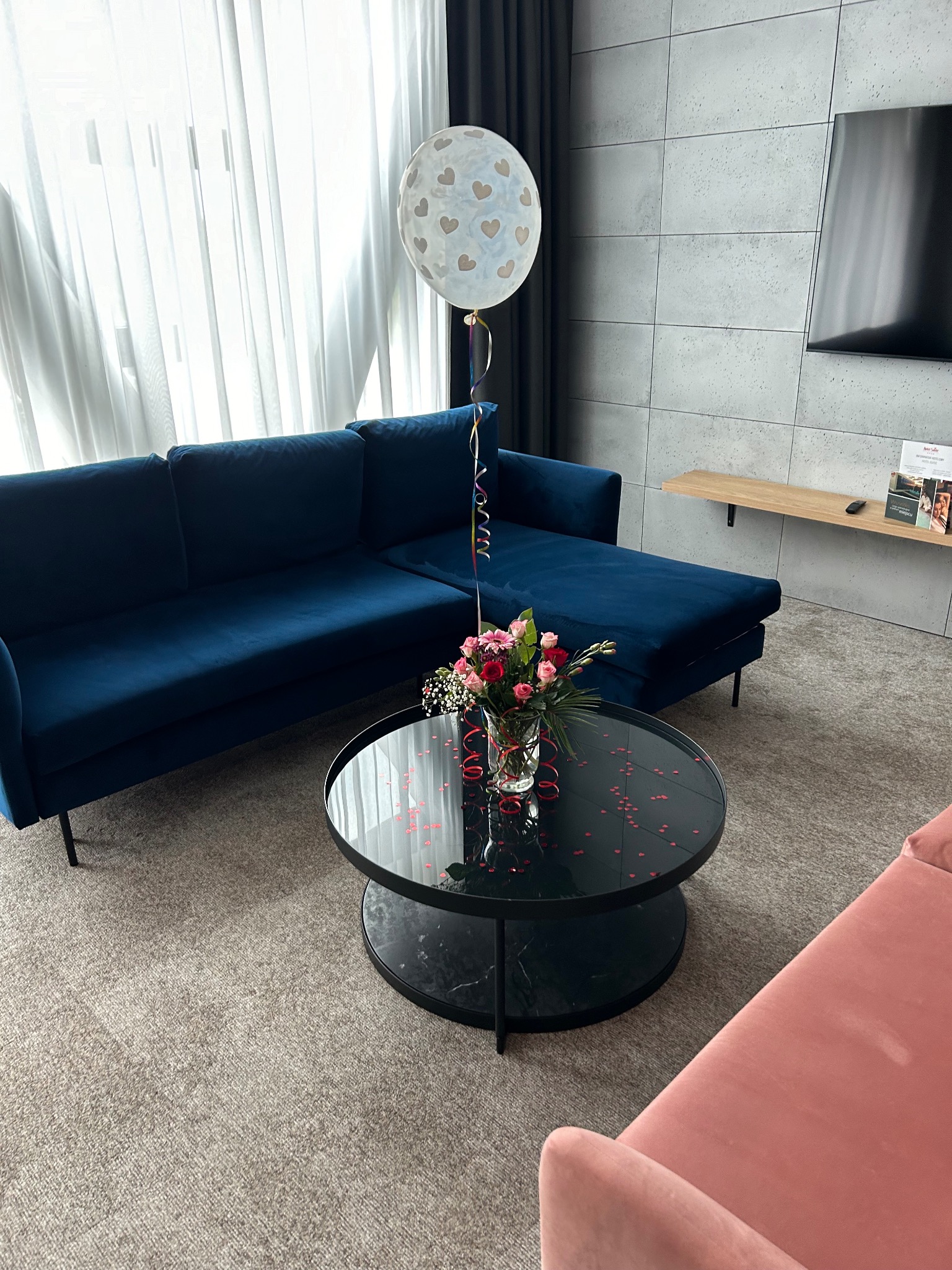 Udekorowany salon z ciemnoniebieską sofą narożną, czarnym stolikiem z kwiatami i balonem. Konfetti w kształcie serc na stoliku. Stonowana kolorystyka wnętrza.