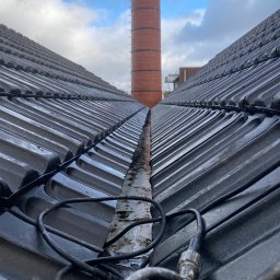 SUN-BIT Spółka z ograniczoną odpowiedzialnością - Dachówka ceramiczna w perspektywie z widocznym kominem w tle i kablem na pierwszym planie, sugerującym prace konserwacyjne lub inspekcję.