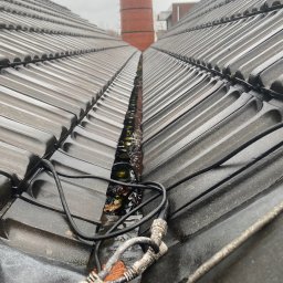 SUN-BIT Spółka z ograniczoną odpowiedzialnością - Mokry dach z dachówki ceramicznej z widocznymi kablami i zanieczyszczeniami w rynnie dachowej, komin w tle.