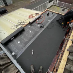 Praca na dachu: dekarz montuje papę termozgrzewalną, używając opalarki. Widoczne narzędzia, częściowo zdjęte dachówki i rolka papy Royal na płaskim dachu.