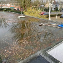 SUN-BIT Spółka z ograniczoną odpowiedzialnością - Zalegająca woda z liśćmi na płaskim dachu w Pabianicach. Widoczne narzędzia do czyszczenia i fragmenty budynków w tle. Dach wymaga interwencji dekarskiej.