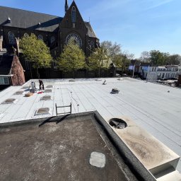 SUN-BIT Spółka z ograniczoną odpowiedzialnością - Praca na dachu: dekarz przy świetlikach na jasnoszarym, płaskim dachu z widokiem na kościół w tle. Widoczne narzędzia i materiały dekarskie.