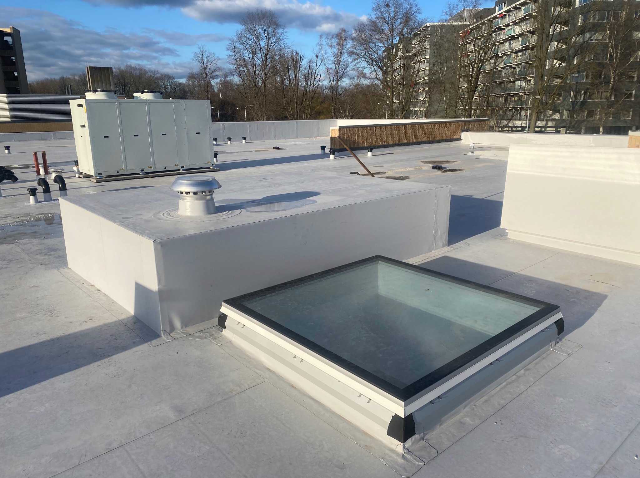 Płaski dach z lukarną i wentylacją, otoczony budynkami. Widoczne detale wykończenia dachu i elementy instalacji wentylacyjnej. Jasne, naturalne oświetlenie.