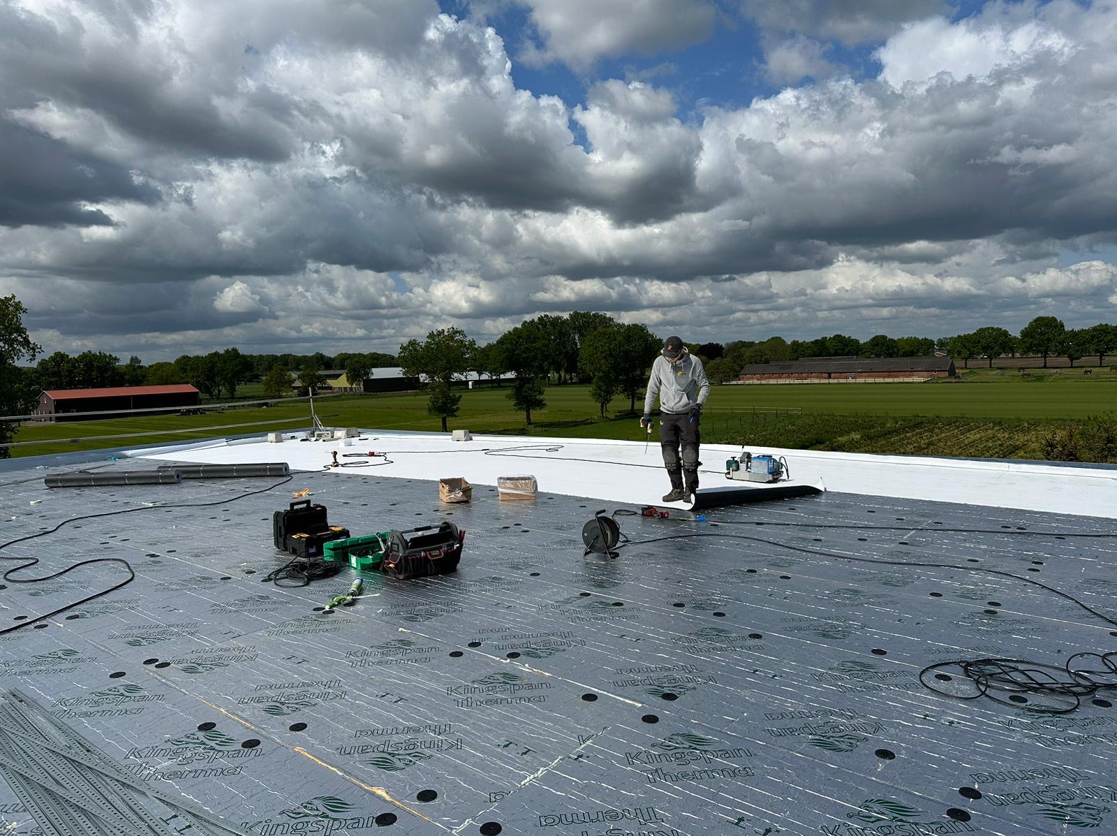 Pracownik w trakcie montażu izolacji Kingspan Therma na dachu. Widoczne narzędzia, rolka papy i elementy mocujące. W tle zielony krajobraz pod błękitnym niebem.