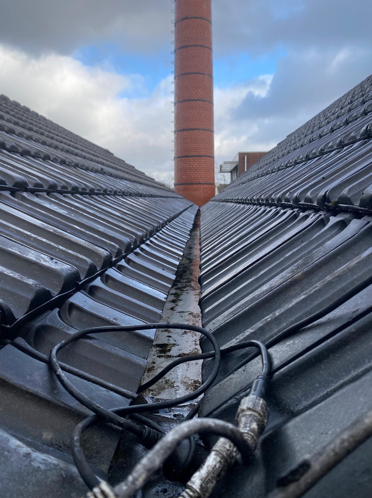 Dachówka ceramiczna w perspektywie z widocznym kominem w tle i kablem na pierwszym planie, sugerującym prace konserwacyjne lub inspekcję.