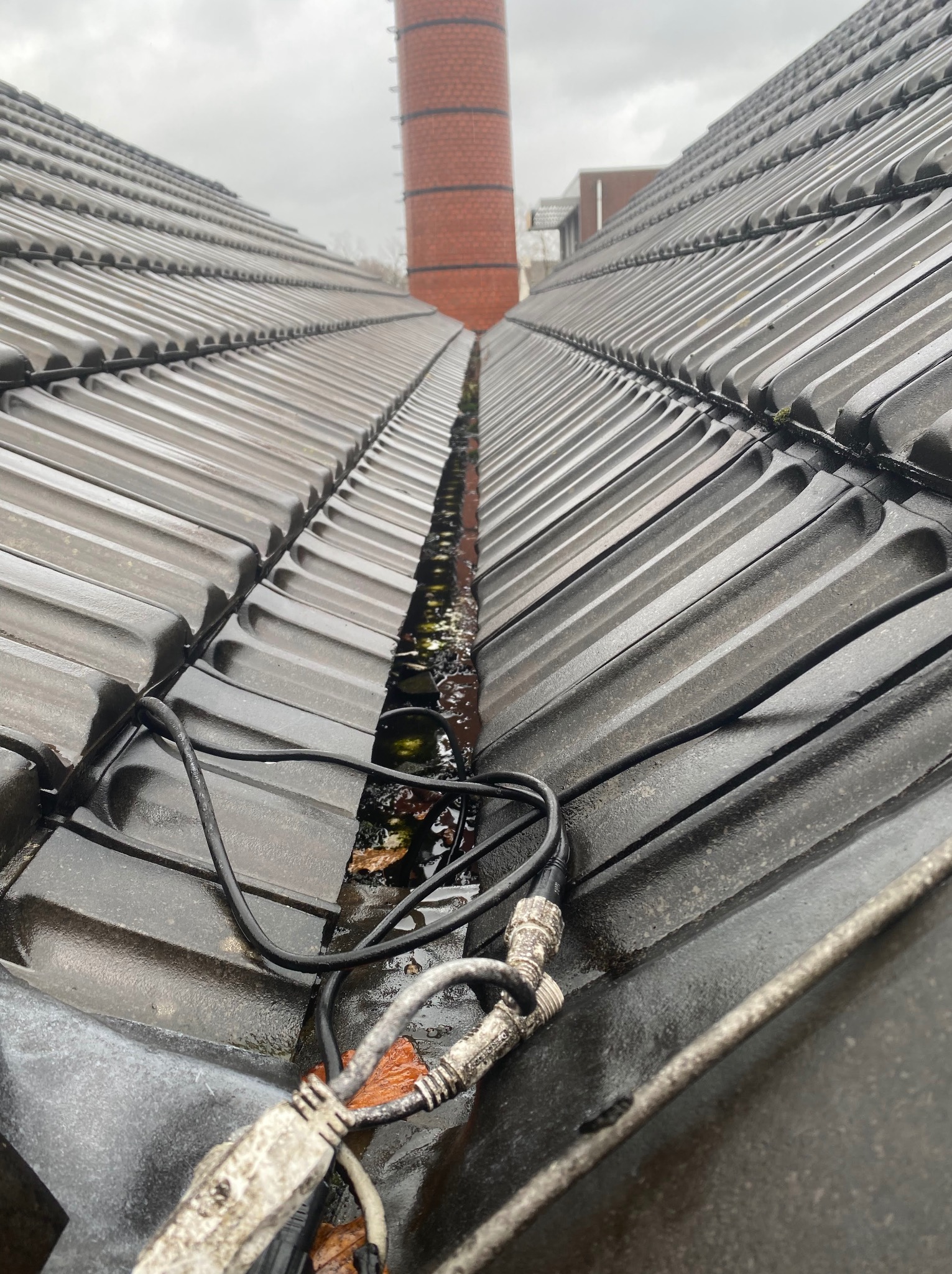 Mokry dach z dachówki ceramicznej z widocznymi kablami i zanieczyszczeniami w rynnie dachowej, komin w tle.