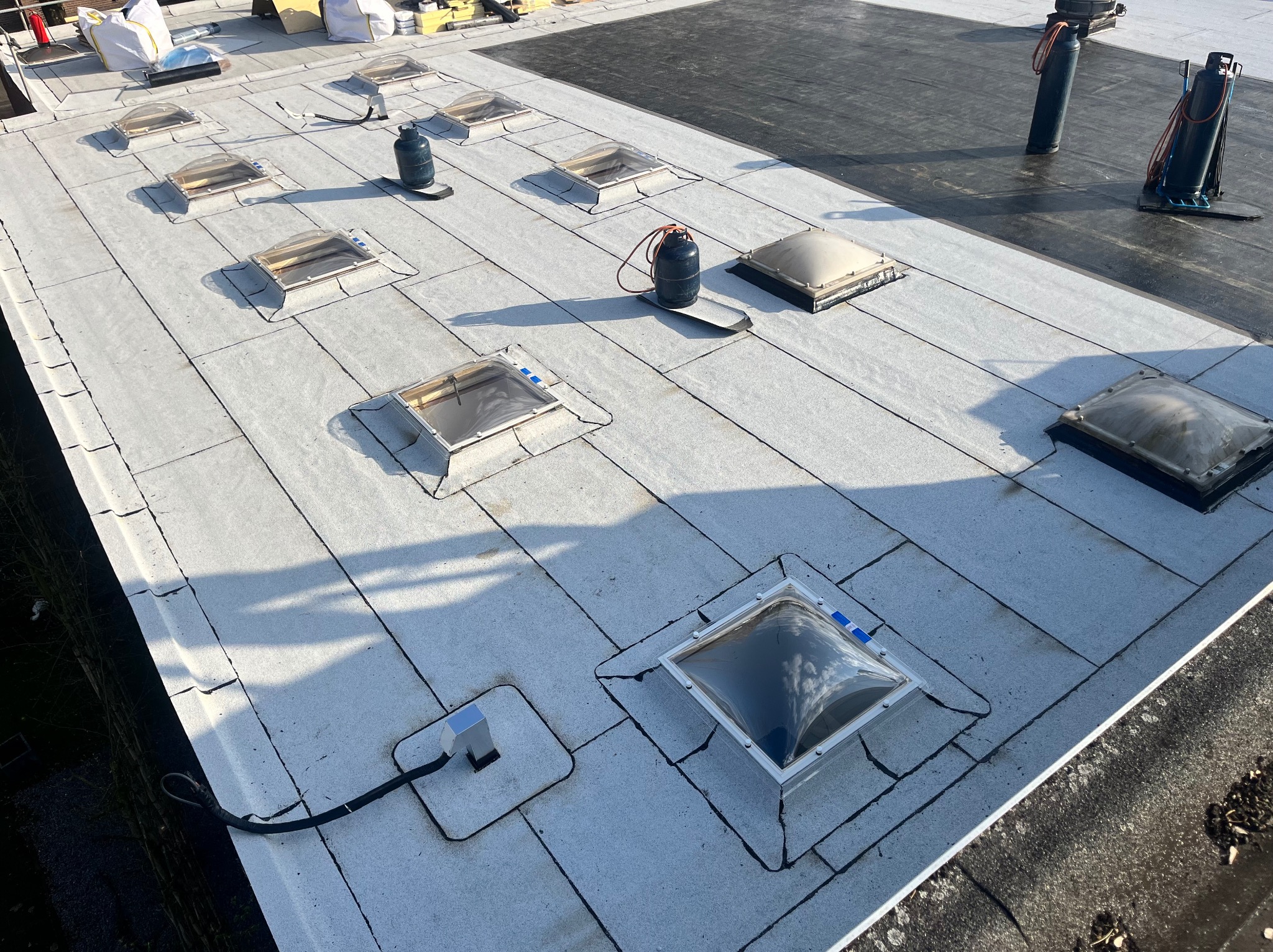 Płaski dach pokryty jasną papą z widocznymi świetlikami i sprzętem dekarskim. Na papie widoczne regularne podziały. Ujęcie z góry, lekko z ukosa.